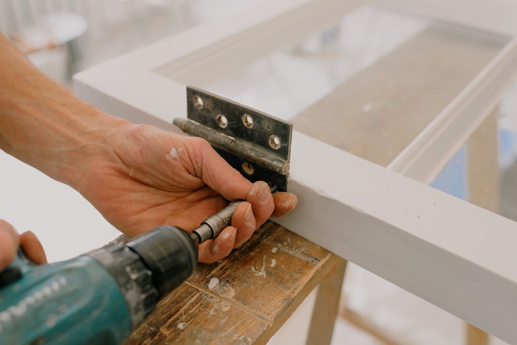 Crop unrecognizable worker drilling metal hinge on white window frame during house renovation